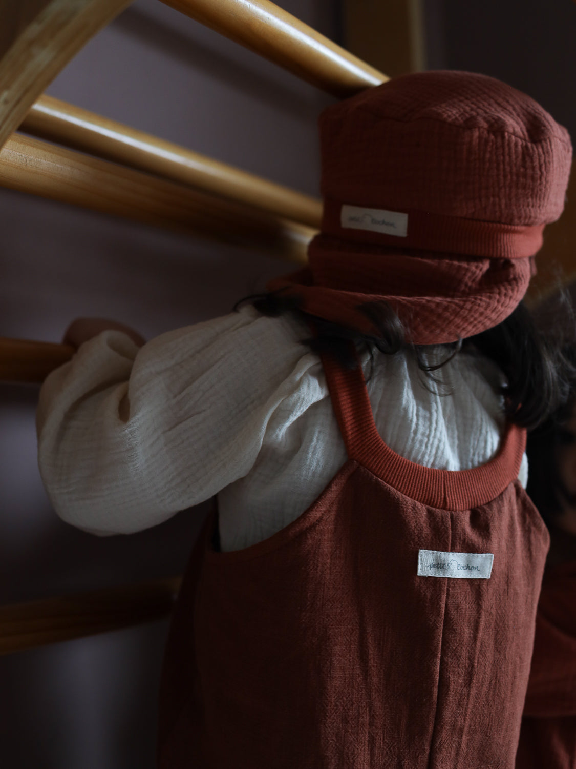 Ein Kleinkind erklimmt eine Kletterstruktur und trägt die Michelmütze Bio-Musselin mit Nackenschutz - Terrakotta, eine luftige Sommermütze aus Bio-Musselin mit elastischem Bündchen, passend zur rostrotfarbenen Kleidung.