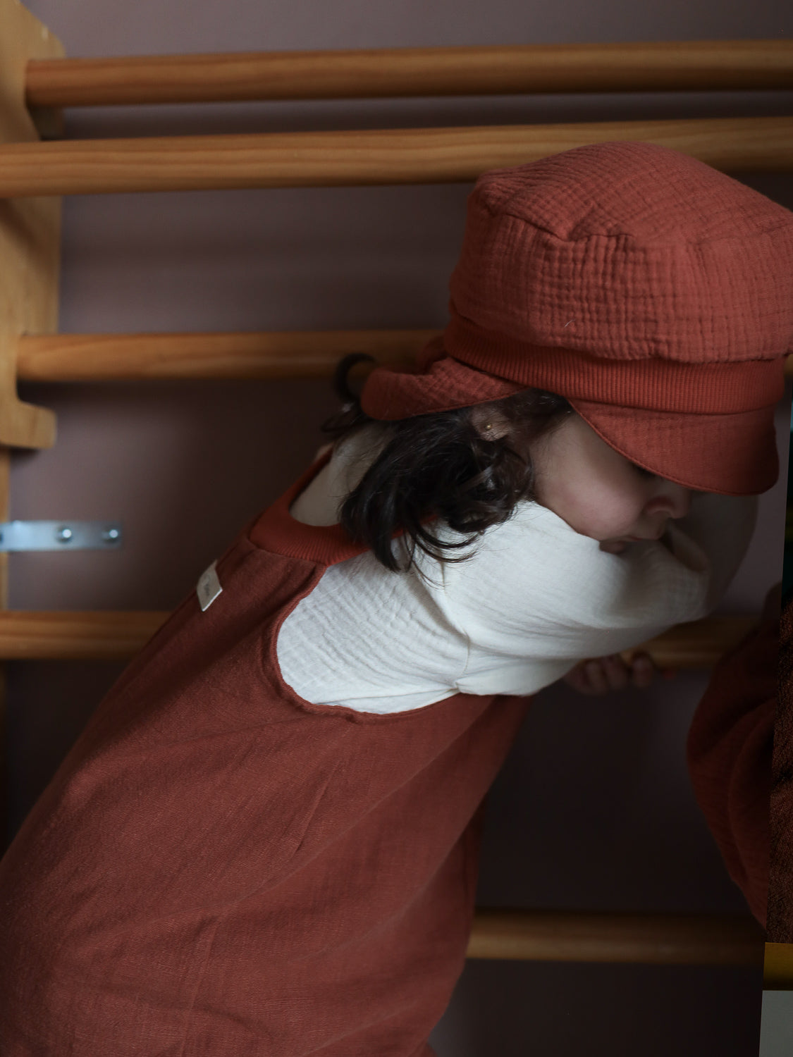 Kleinkind an Kletterwand, trägt die Michelmütze Bio-Musselin mit Nackenschutz - Terrakotta; luftige Sommermütze aus Bio-Baumwolle mit elastischem Bündchen, schützt den Kopf und wächst mit. Gesicht teils verdeckt.