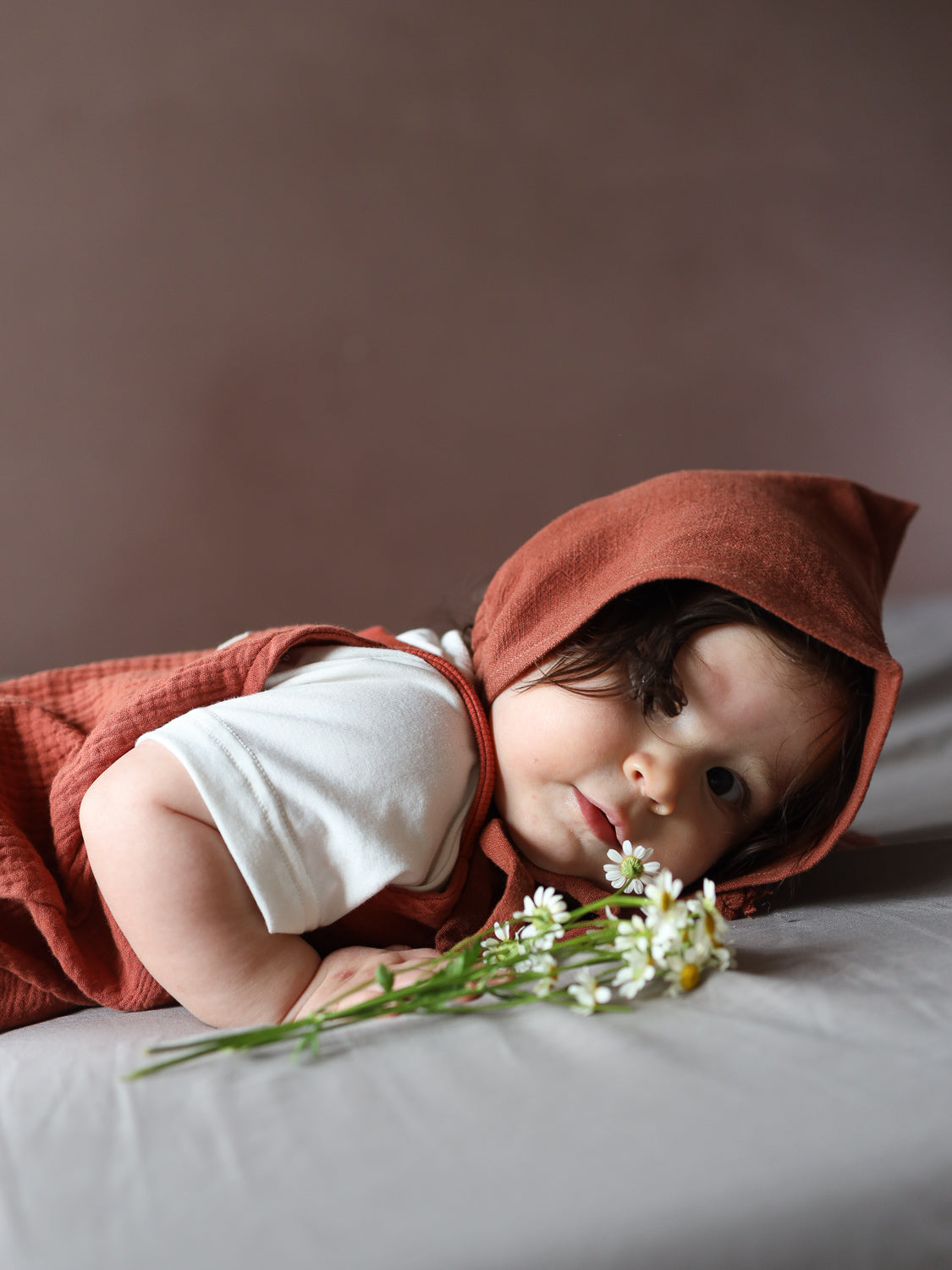 Baby in einem rostfarbenen Zwergenmütze Leinen - Ziegel, liegend auf dem Bauch. Die Mütze hat eine spitze Form, die Kopf und Ohren schützt. Das Baby greift nach weißen Blumen.
