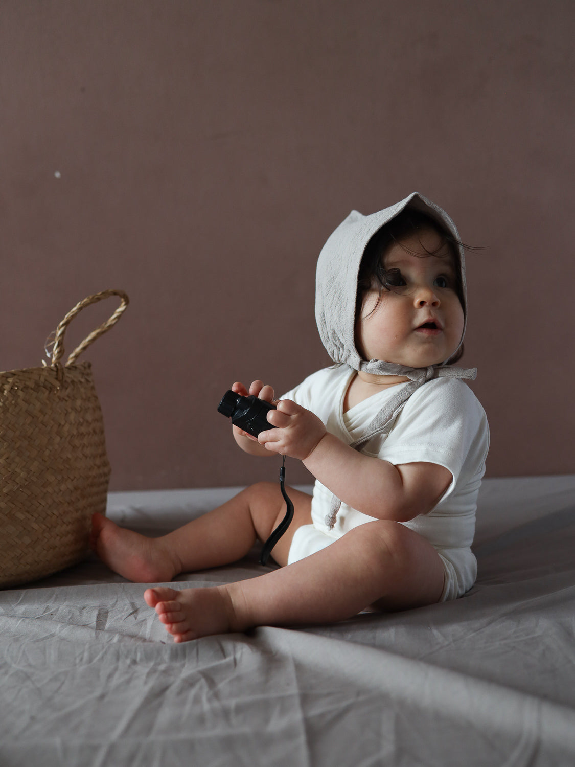 Ein Baby trägt die Zwergenmütze Leinen - Sand, gebunden unter dem Kinn, und hält neugierig ein kleines schwarzes Objekt. Neben ihm steht ein geflochtener Korb.