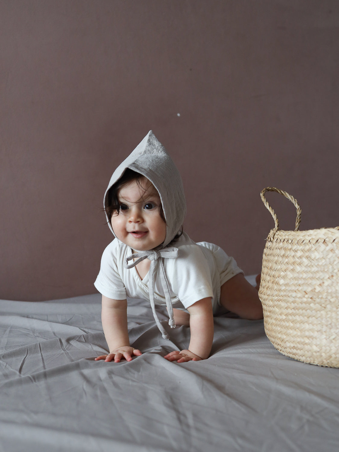 Ein Baby trägt die Zwergenmütze Leinen - Sand, eine spitze, gebundene Haube, während es auf einem grauen Laken krabbelt. Daneben steht ein geflochtener Korb.
