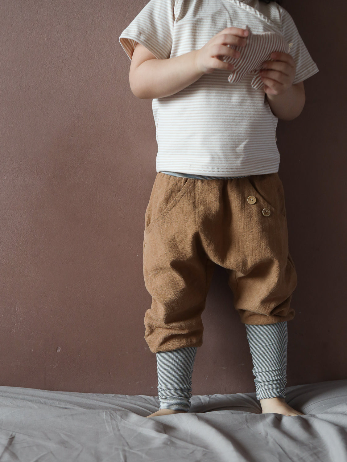 Ein Kleinkind trägt das Kurzes Wickelshirt Baumwolle - Ringel Sand, kombiniert mit lockeren braunen Hosen und gestreiften Leggings. Es hält ein gestreiftes Stofftier, stehend auf einer weichen Oberfläche.
