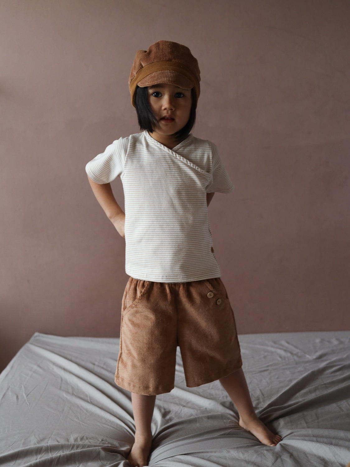 Ein Kind trägt das Kurzes Wickelshirt Baumwolle - Ringel Sand, ein gestreiftes, bequemes Oberteil mit Wickelausschnitt, kombiniert mit braunen Cord-Shorts und passender Mütze, stehend auf grauem Stoff.