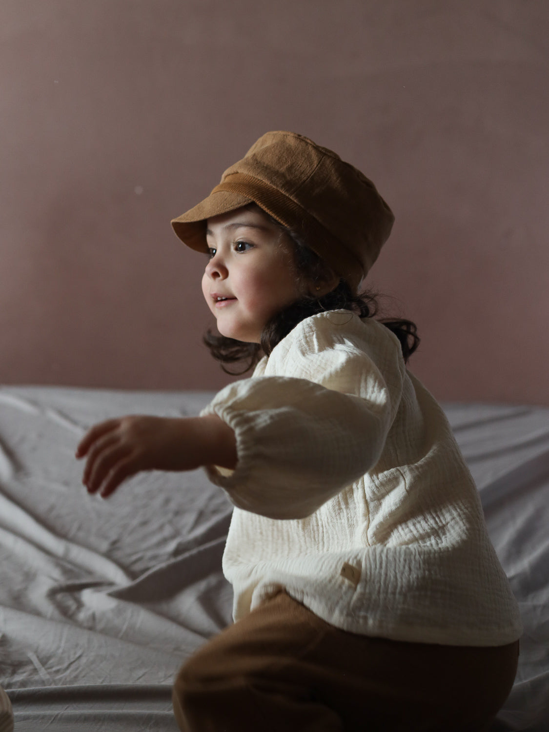 Ein Kleinkind trägt die Michelmütze Leinen - Weizen, zusammen mit einer cremefarbenen, strukturierten Langarmbluse und braunen Hosen, während es auf einer weichen Unterlage sitzt und neugierig nach vorne schaut.
