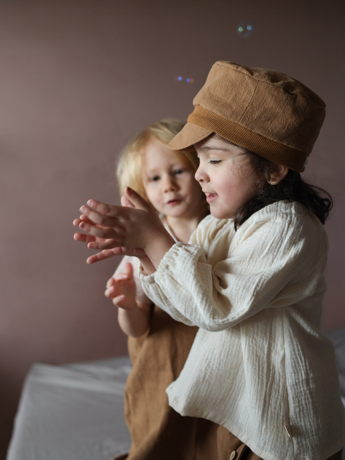 Zwei Kinder spielen drinnen. Im Vordergrund trägt ein Mädchen eine Michelmütze Leinen - Weizen und klatscht in die Hände. Ein weiteres Kleinkind beobachtet sie. Seifenblasen schweben um sie herum.