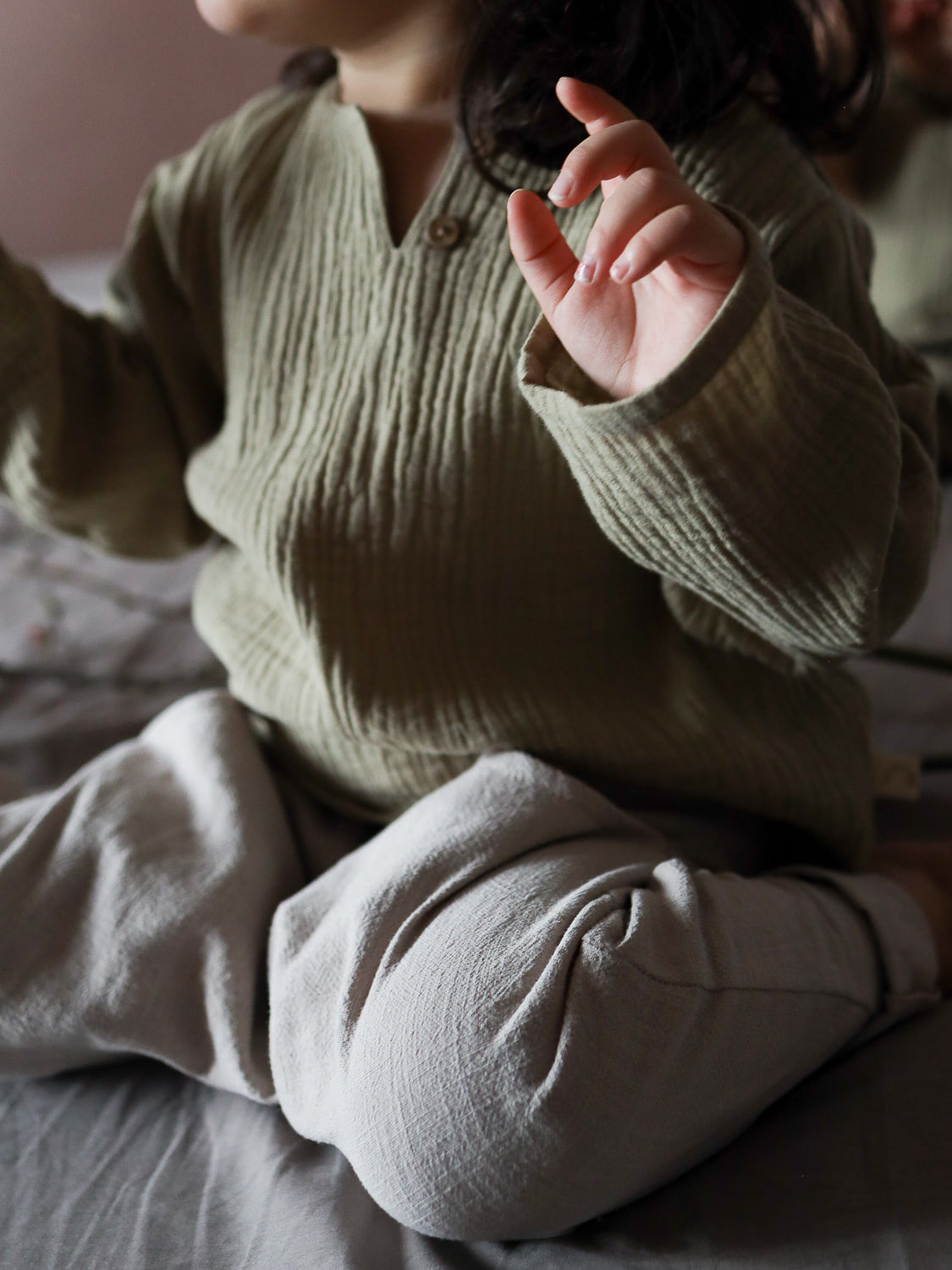 Ein Kind trägt das Hemdchen Musselin - Salbei, ein leichtes, langärmeliges Oberteil aus Bio-Musselin mit strukturiertem Stoff, ideal für warme Tage. Das Bild zeigt den Oberkörper und sanft erhobene Hände.