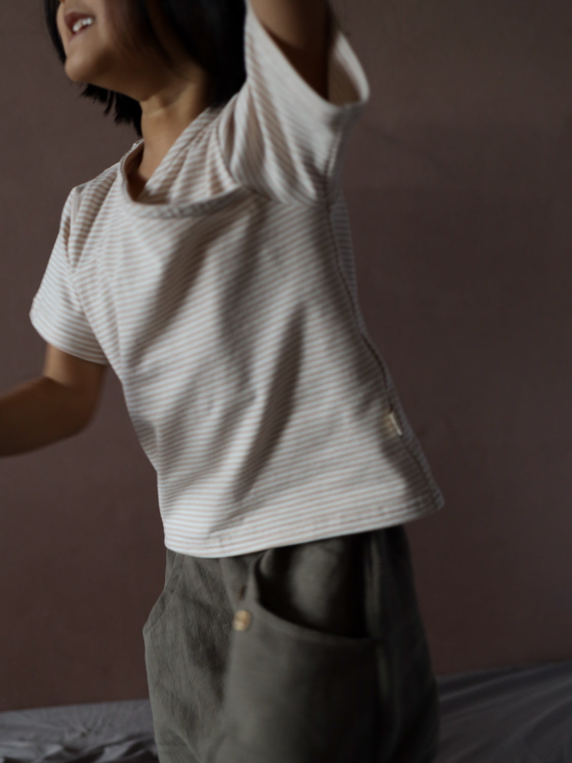 Ein Kind trägt ein Kurzes Wickelshirt Baumwolle - Ringel Sand mit schmalen Streifen, kombiniert mit locker sitzenden olivgrünen Hosen. Der weiche Stoff des Shirts wirkt bequem und modisch zeitlos.