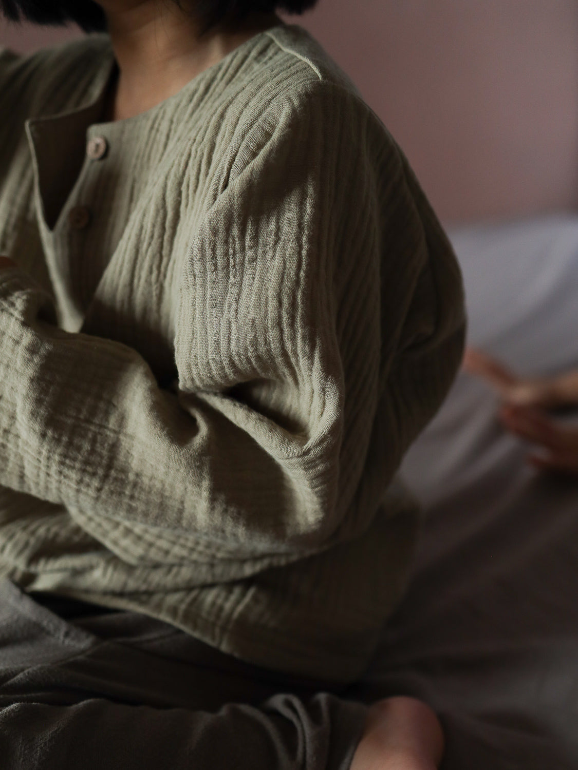 Nahaufnahme einer Person mit einem entspannten Sitz, die ein weiches, langärmeliges Hemdchen Musselin - Salbei trägt, mit Knöpfen am Ausschnitt und sichtbarer Stoffstruktur, ideal für Babys und Kleinkinder.