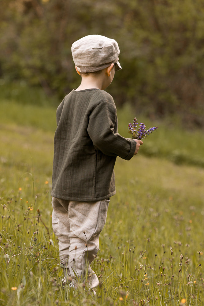 Ein Kind trägt die Michelmütze Leinen - Sand, passend zu seiner Kleidung, während es einen Strauß lila Wildblumen hält. Die Mütze ergänzt den natürlichen Look im Hintergrund der Wiese.