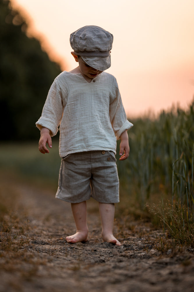 Ein Kleinkind trägt die Michelmütze Leinen - Sand, während es barfuß auf einem schmalen Pfad geht, gekleidet in einem locker sitzenden, langärmeligen Leinenshirt und neutralen Shorts, umgeben von hohen Gräsern.
