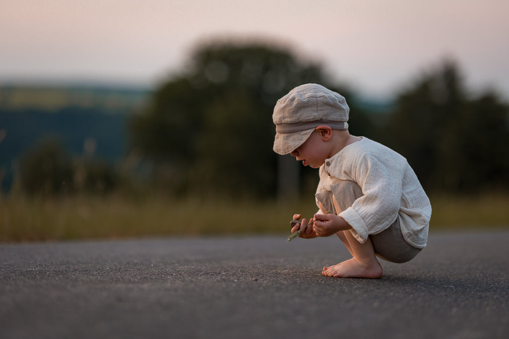 Ein kleines Kind, barfuß und in lockerer Leinenkleidung, hockt auf einer Straße und hält sanft eine Blumenranke. Es trägt die Michelmütze Leinen - Sand, passend zur ruhigen, ländlichen Umgebung.