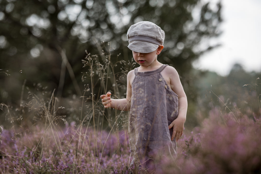 Ein Kleinkind steht in einem Feld, trägt die Michelmütze Leinen - Sand, eine weiche Kappe über der Stirn, kombiniert mit lockeren Leinenoveralls in einer ruhigen, natürlichen Umgebung.