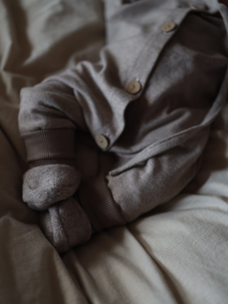 Unterer Teil eines Babys in einem weichen, grauen Mini Overall Feinstrick - Hafer Meliert mit Holzknöpfen, passenden Booties, auf einer leicht zerknitterten Decke liegend. Perfekt für Komfort und Bewegungsfreiheit.