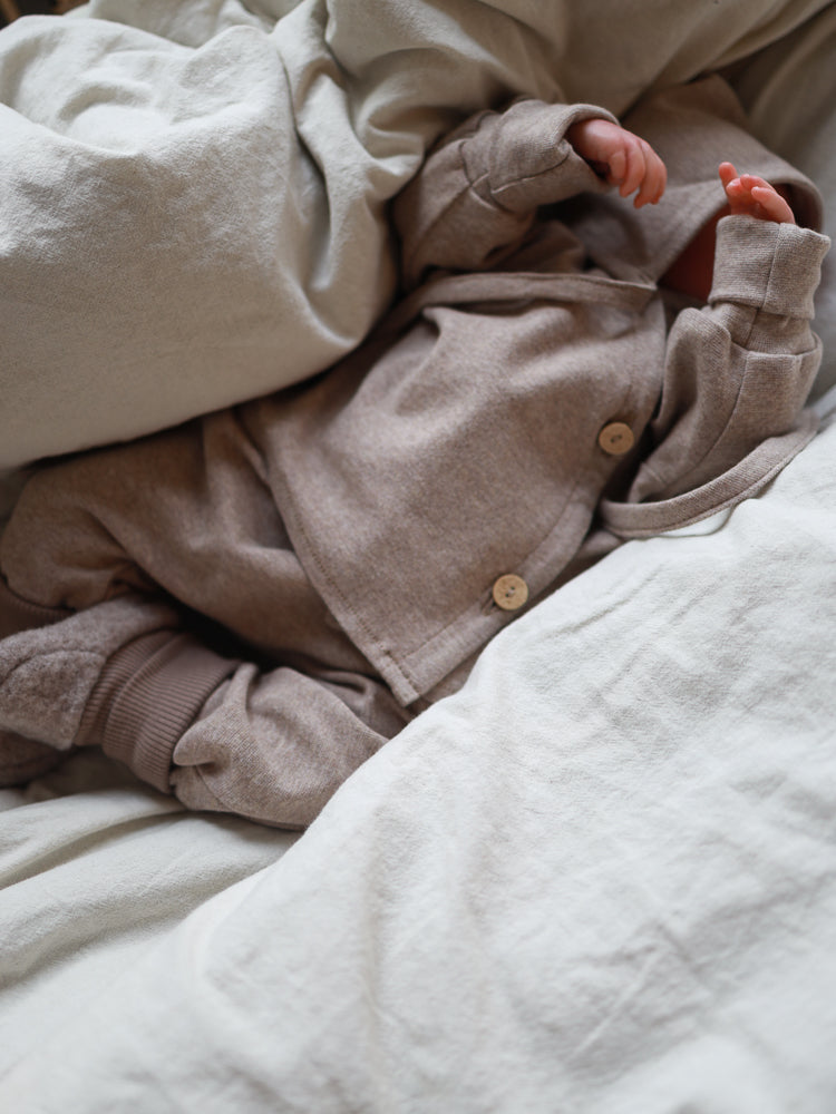 Ein Baby liegt auf einem Bett, teils bedeckt mit neutraler Bettwäsche, trägt den Babyschlüttli Feinstrick - Hafer Meliert. Das Outfit ist beige, zeigt Knopfdetails und kleine Hände und Füße.