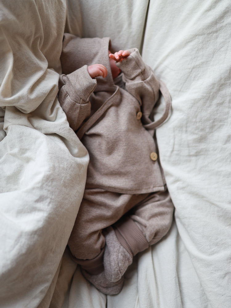 Nouveau-né sur un tissu doux, portant l’ensemble pour bébé en tricot fin – avoine chiné : veste à capuche avec boutons en bois, pantalon assorti et jambières. Mains et jambes visibles dans un environnement naturel et apaisant.