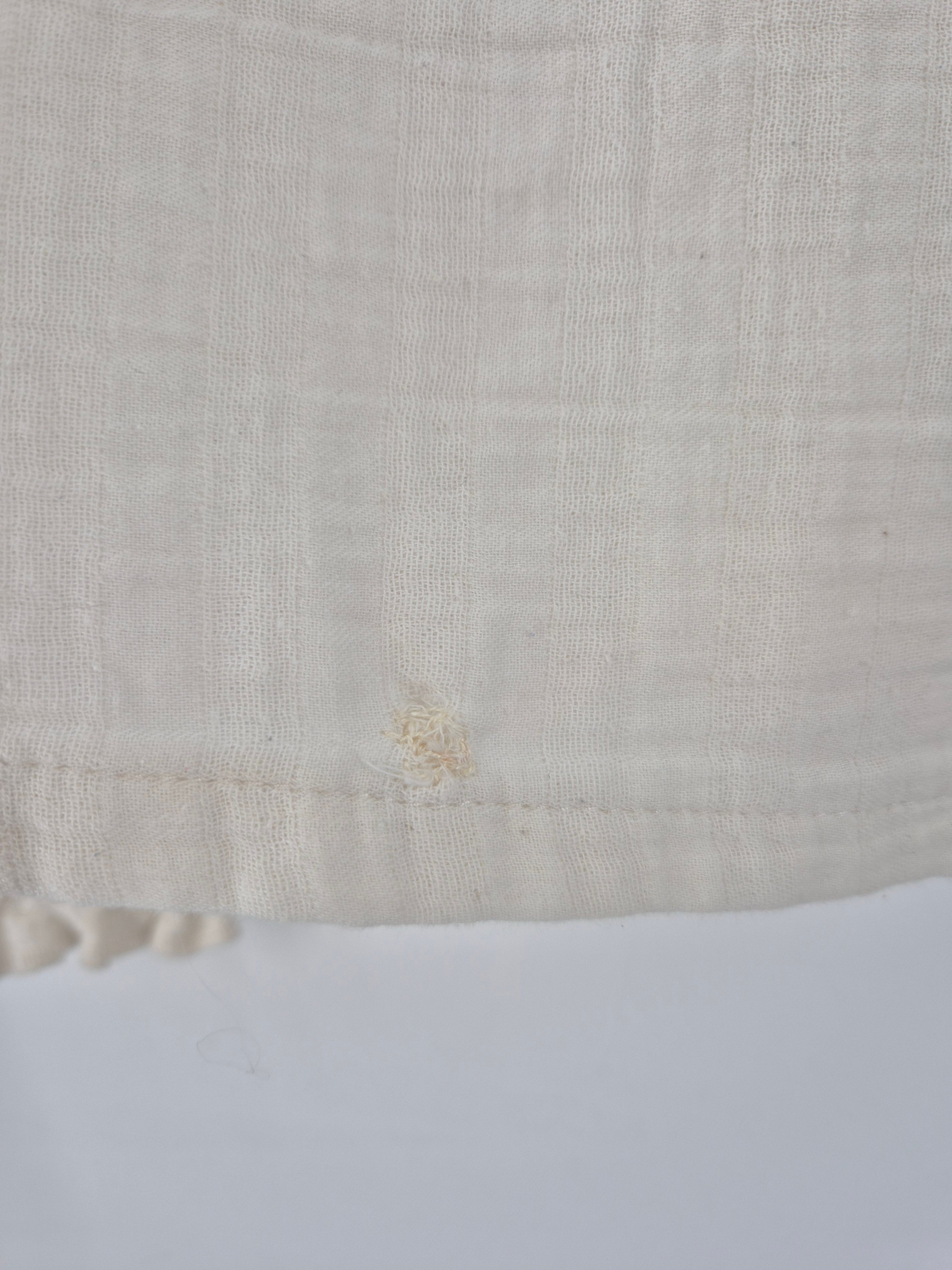 Nahaufnahme der Bluse Bio Musselin Natur: Locker gewebter, off-weißer Stoff mit sichtbarem Gittermuster, gesäumtem Rand und kleinem Knoten durch gezogene Fäden, Zustand gut, leichte Verfärbungen und Flickstellen sichtbar.