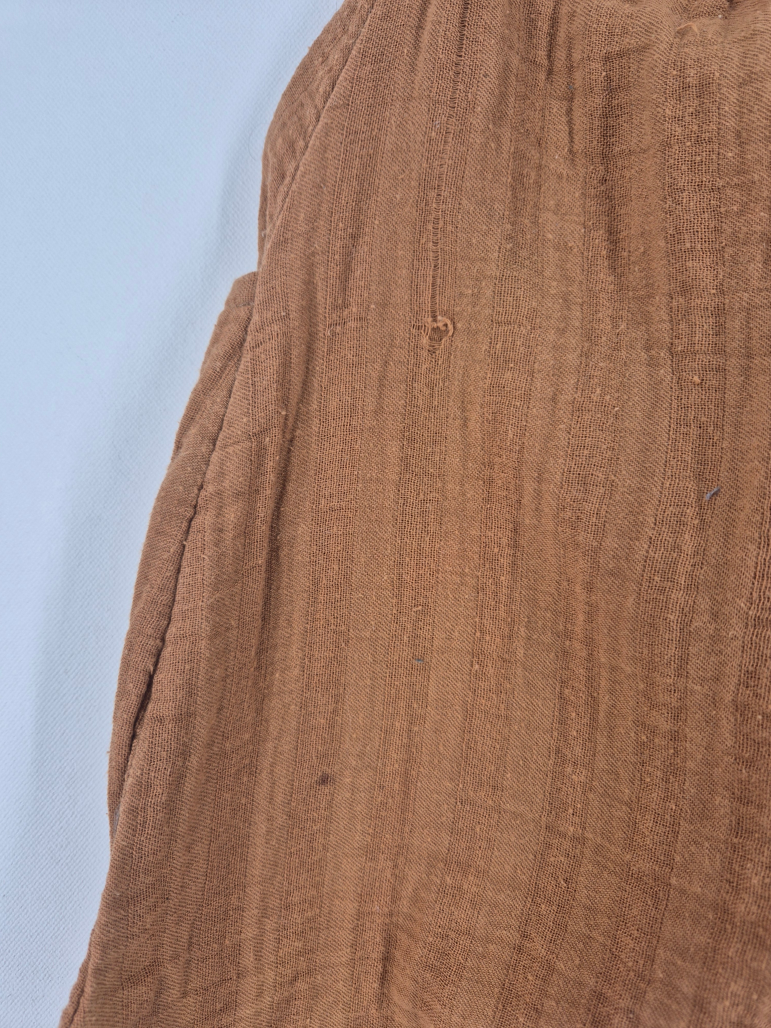 Sommerknicker Bio Musselin Sahara Gr. 3Y aus rostbraunem, leichtem Gauze-Stoff mit seitlicher Tasche. Zeigt einen kleinen Riss vorne am Knie, auf hellem Hintergrund drapiert.