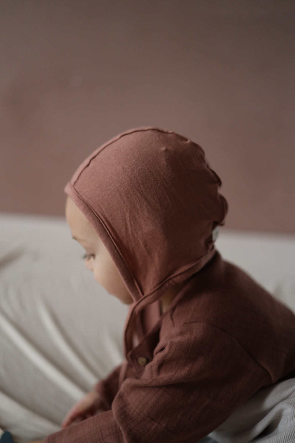 Baby trägt das Babyhäubchen Kaschmir-Mix - Zimt, kuschelweich und elastisch, idealer Schutz für den Kopf, passend zu Loopbody und Babyhose.