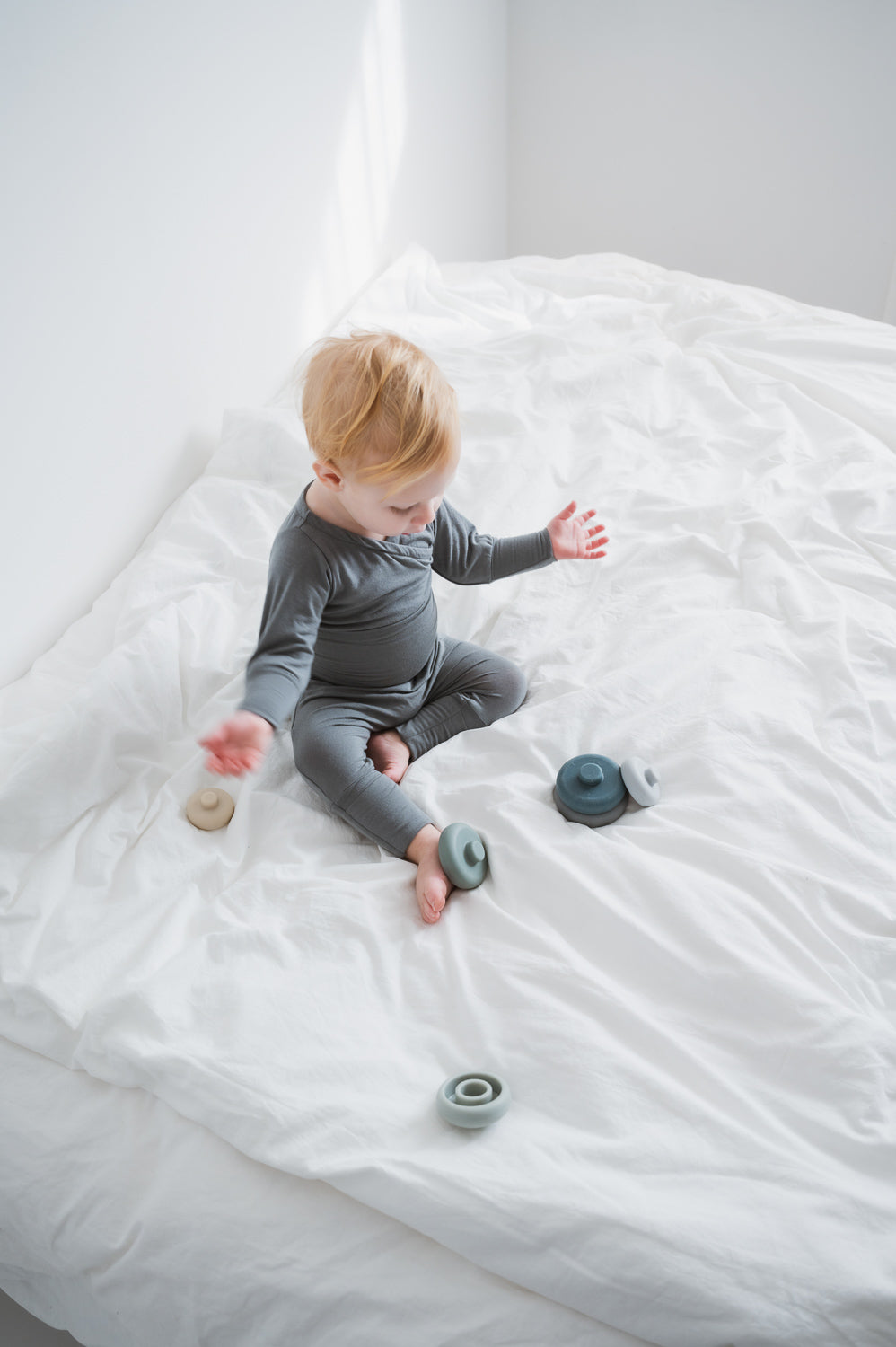 Baby auf Bett spielt mit Spielzeug, trägt Babyknicker Kaschmir-Mix mit Klappbündchen, passend zum Loopbody. Die Hose bietet weiches Material und mitwachsende Passform, ideal für Neugeborene. Erstausstattung Baby und Geschenk für Babys.