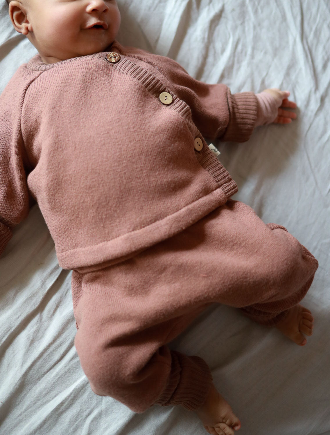 Ein Baby trägt das Babyschlüttli Strick - Nougat aus recycelter Baumwolle mit Krempelbündchen, auf einem Bett liegend.
Erstausstattung Babys. Perfekte Baby Geschenke.