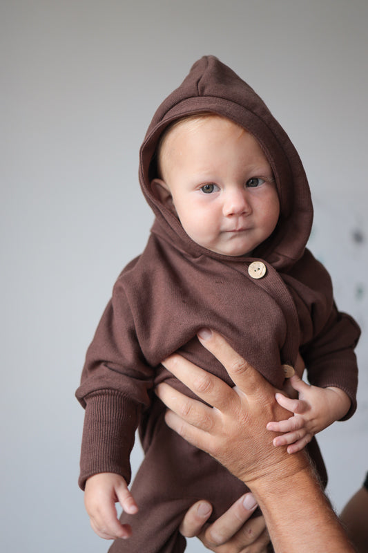 Ein Baby trägt den Kuschelanzug Bio-Sweat - Schoko mit Wichtelkapuze, während es von einer Hand gehalten wird. Der Anzug wirkt kuschelig und warm, ideal für verschiedene Jahreszeiten.  Erstausstattung Baby und Baby Geschenk zur Geburt.