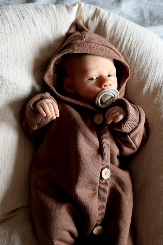 Baby trägt einen Kuschelanzug Bio-Sweat - Schoko mit Wichtelkapuze. Der Overall ist weich angeraut und bietet Komfort und Wärme, ideal für drinnen und draußen.  Erstausstattung Baby und Baby Geschenk zur Geburt.
