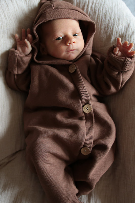 Baby in einem Kuschelanzug aus Bio-Sweat mit Wichtelkapuze, liegend auf einer Decke. Der Overall ist großzügig geschnitten und hat Krempelbündchen, perfekt für verschiedene Jahreszeiten.  Erstausstattung Baby und Baby Geschenk zur Geburt.