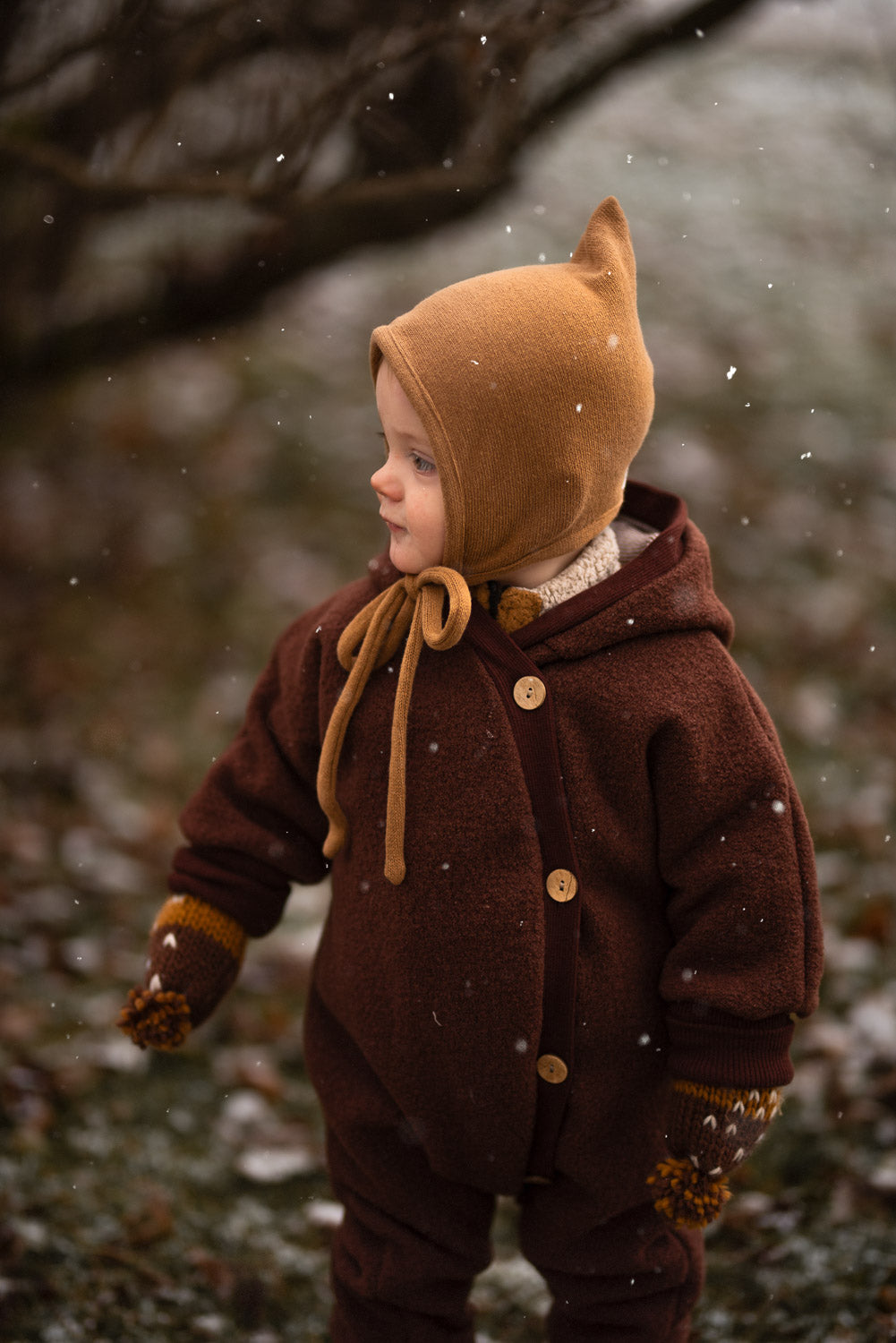 Niño con mono de lana afieltrada color castaño y gorro de punto con diseño de duendes, ideal para las estaciones frescas. El mono ofrece regulación térmica y libertad de movimiento, perfecto para actividades al aire libre.