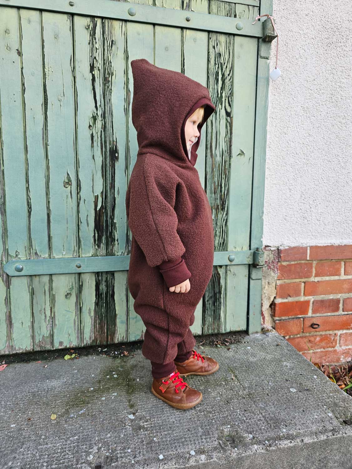 Kind in einem Kuschelanzug Wollwalk - Kastanie mit Kapuze, zeigt eng anliegende Bündchen, ideal für kaltes Wetter, atmungsaktiv und wärmeregulierend für optimalen Tragekomfort.