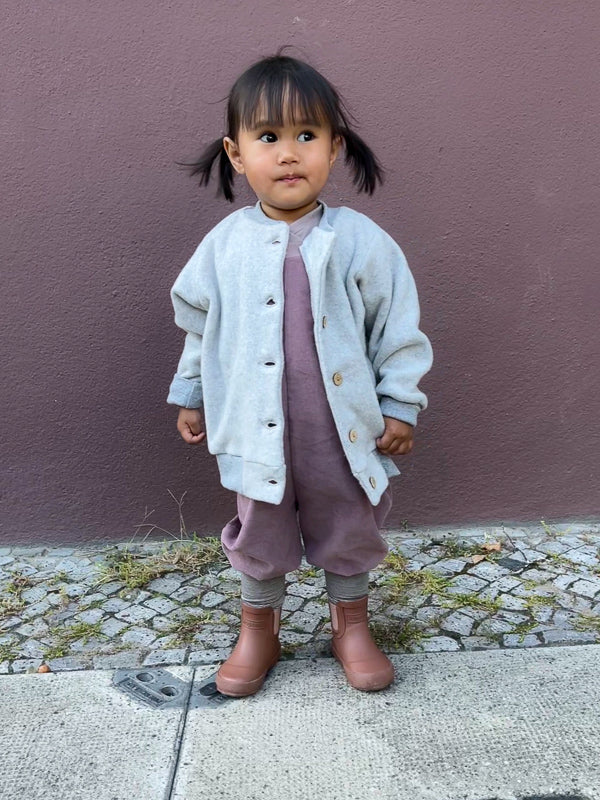 Mädchen trägt Schlupflatzhose Feincord - Malve vor lila Wand. Bequeme, handgenähte Latzhose aus weichem Cord, ideal für Kinder in verschiedenen Altersstufen.