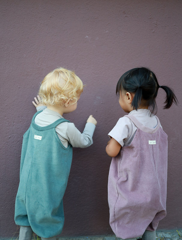 Zwei Kinder in Schlupflatzhose Feincord - Malve, eine weiche Baumwollhose ohne komplizierte Verschlüsse, stehen vor einer Wand. Perfekt für einfaches Anziehen, ideal für Kleinkinder. nachhaltige Kinderkleidung made in Berlin