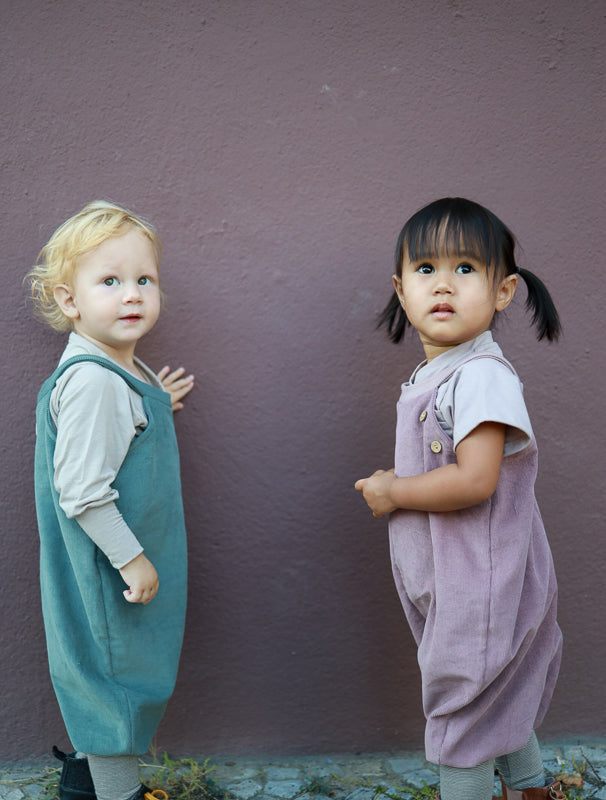 Zwei Kinder stehen nebeneinander, eines trägt die Schlupflatzhose Feincord - Malve, eine weiche und bequeme Cord-Latzhose ohne komplizierte Verschlüsse, ideal für einfaches An- und Ausziehen. nachhaltige Kinderkleidung made in Berlin