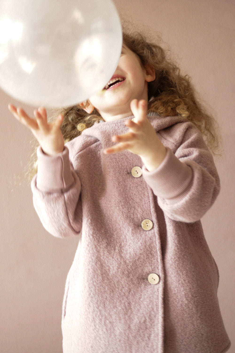 Girl with curly hair in a pink boiled-wool Pixie jacket – Powder, playing with a bubble. Close-up shows the jacket’s buttons and organic jersey lining.