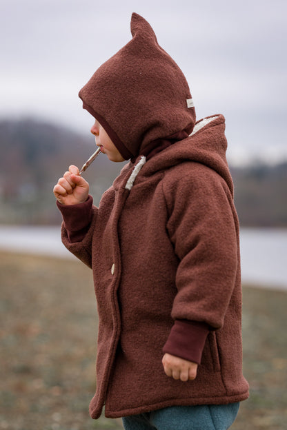 Child wearing the “Wichteljacke” boiled-wool jacket in chestnut, with a hood and roll-up cuffs, ideal for autumn and winter, made from organic materials.
Boiled-wool jacket and winter jacket for children. Hand-sewn in Berlin
